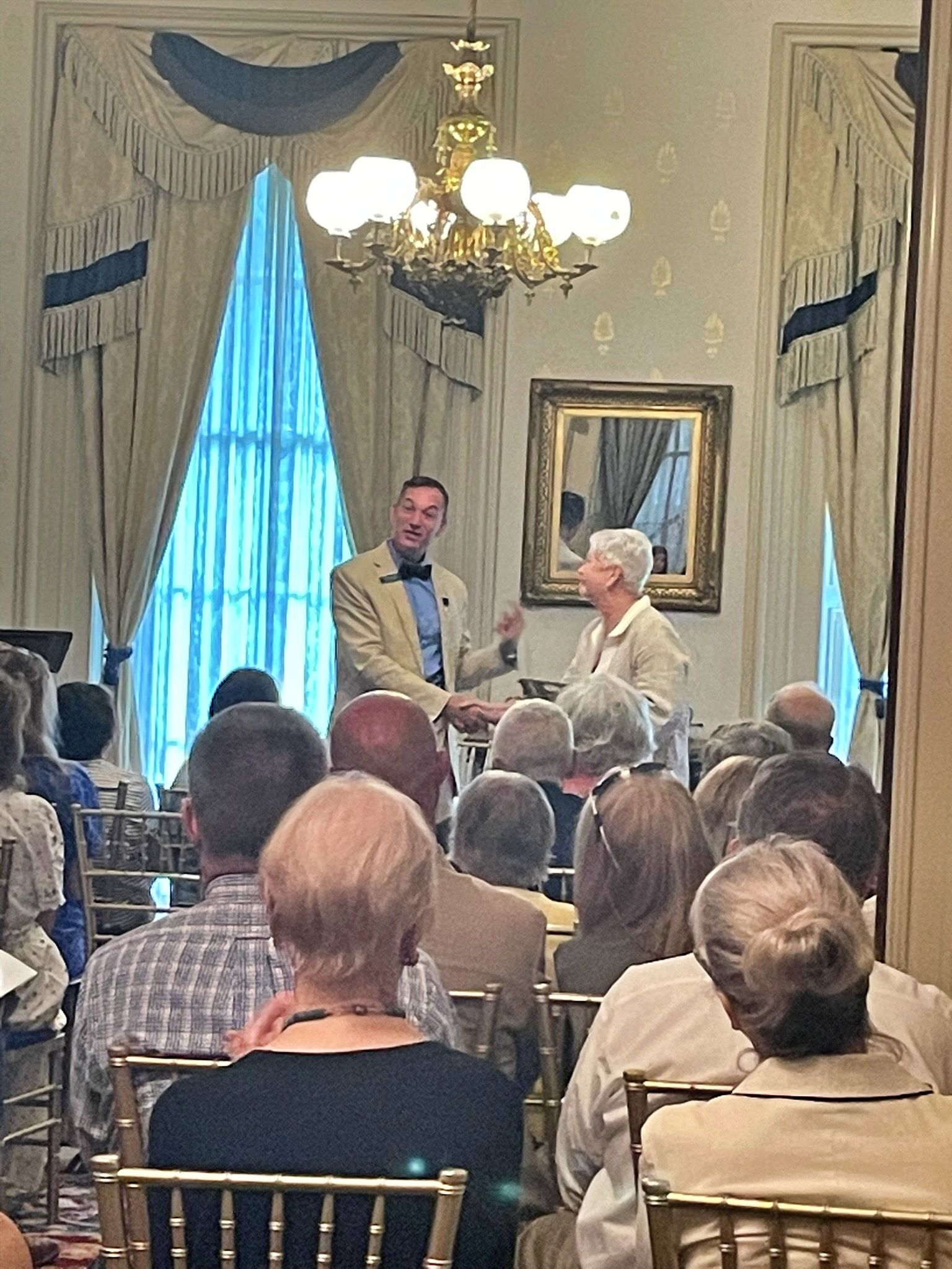 A person in a tan blazer shakes hands with another person in a formal room with a chandelier and large draped windows.
