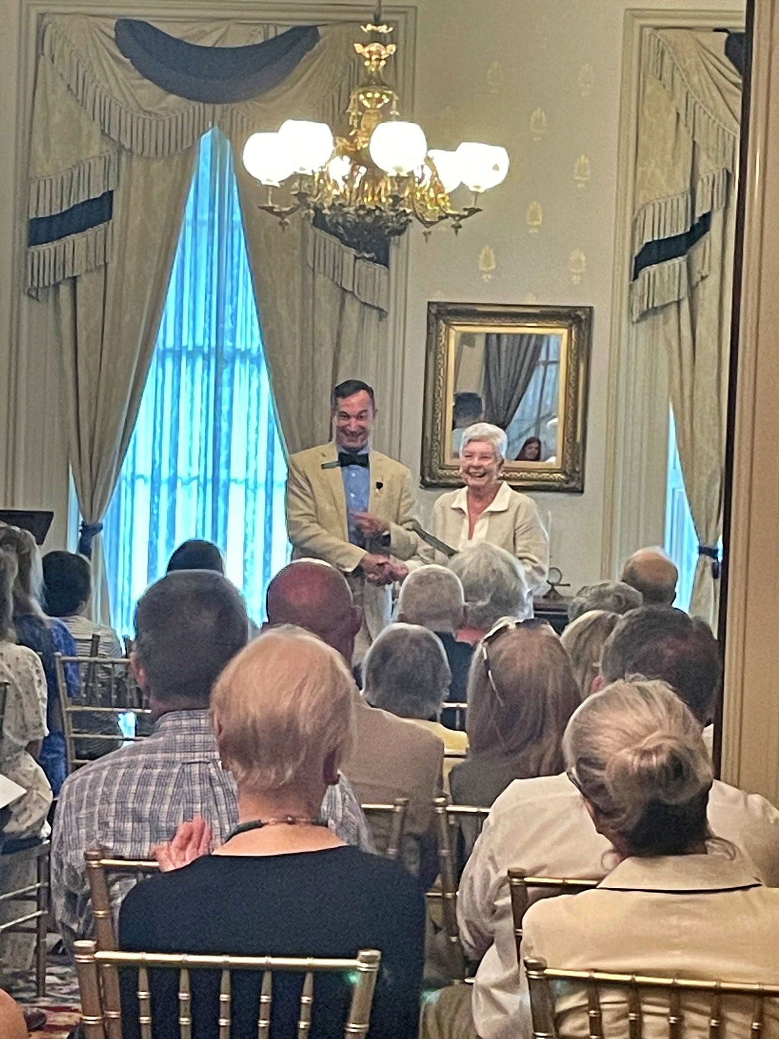 A presenter speaks to an audience in a formal, well-lit room with draped windows, a chandelier, and a framed mirror.