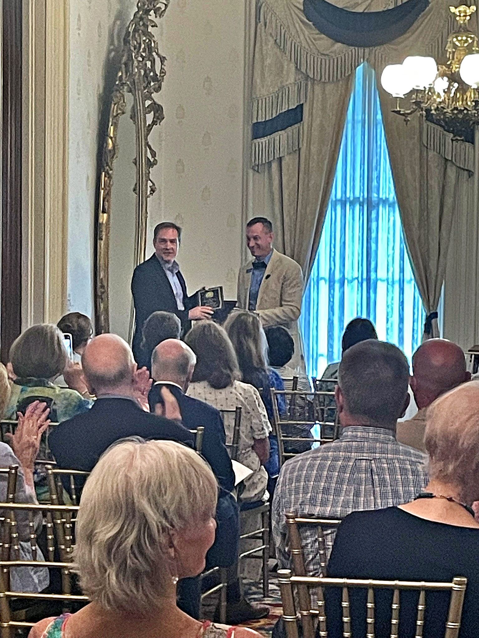 Two men in suits stand in a formal room, one handing an award to the other before an audience seated at round tables.