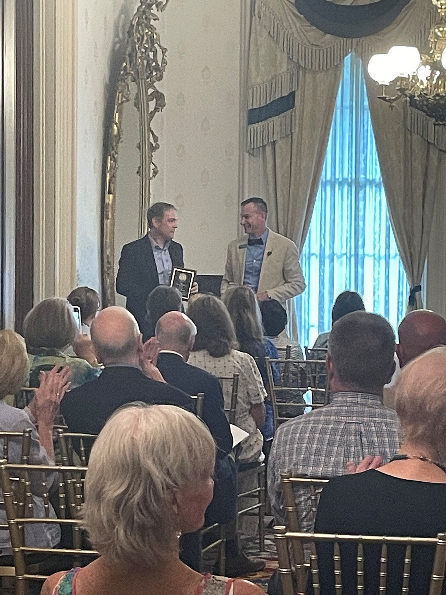 Two men stand at a podium in a formal, sunlit room, addressing an audience seated in rows of gold chairs.