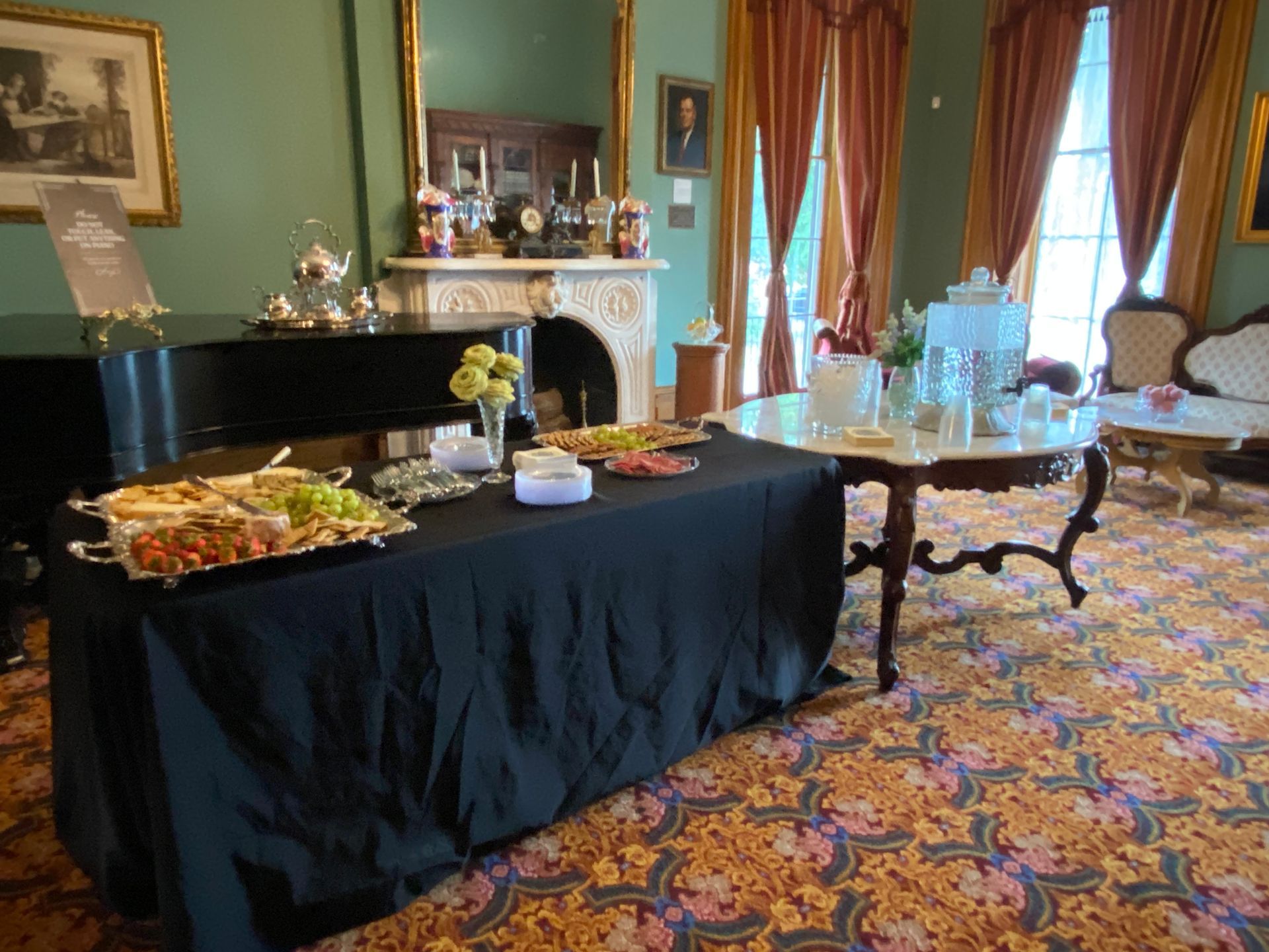 A black-clothed catering buffet table and a round table with drinks set up inside a decorated, ornate reception room.