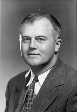 A head-and-shoulders studio portrait of a man in a suit and patterned necktie, set against a plain, neutral background.