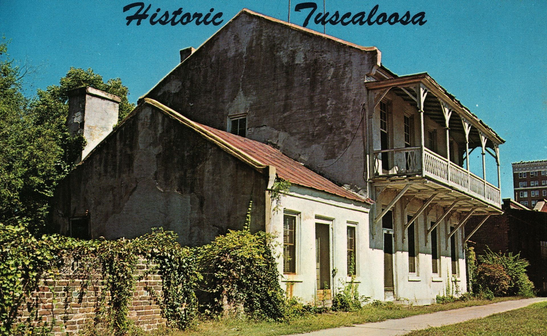 A vintage postcard of a historic, two-story white building in Tuscaloosa with a wooden balcony and brick chimney.