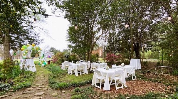 Outdoor garden party setup with white-clothed tables, chairs, and a balloon arch, beneath trees with string lights.
