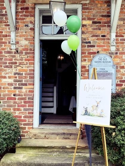 A brick building entrance decorated with green and white balloons, featuring a welcome sign on an easel in the foreground.