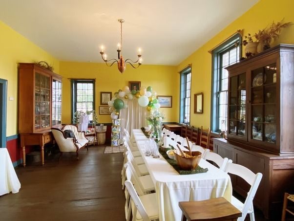 A bright yellow dining room with a long table, white tablecloths, chairs, wooden cabinets, and a festive balloon arch.