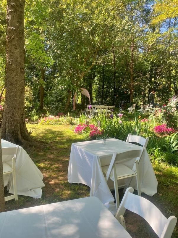 Outdoor tables with white tablecloths and folding chairs set on grass in a garden with trees and flowering plants.