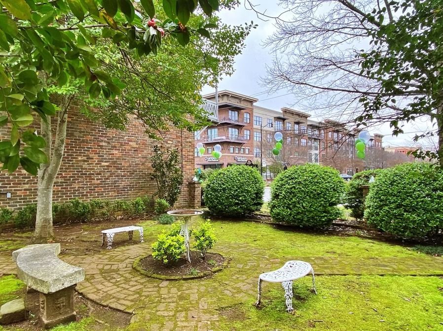 A small garden with stone benches, a central birdbath, and green bushes, with a brick wall and city building in background.