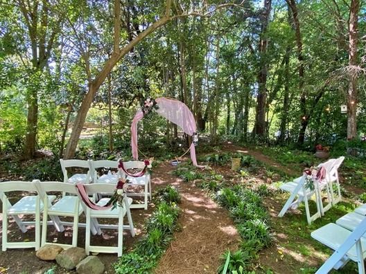 A forest wedding setup with white chairs lining a dirt aisle leading to a pink-draped archway decorated with flowers.