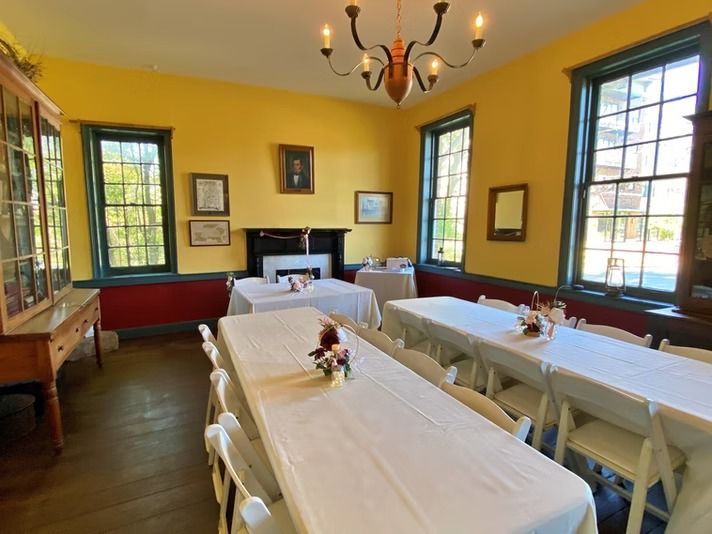 A dining room with yellow walls, a fireplace, and rows of tables covered in white tablecloths set for a gathering.
