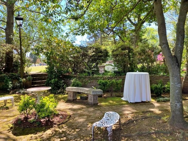 A sunlit garden terrace with a stone bench, a small round table, a cocktail table with a white cloth, and a metal chair.
