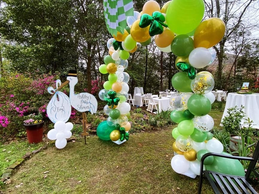 A festive balloon arch in shades of green, yellow, and white stands in a garden near a decorative cutout of a stork.