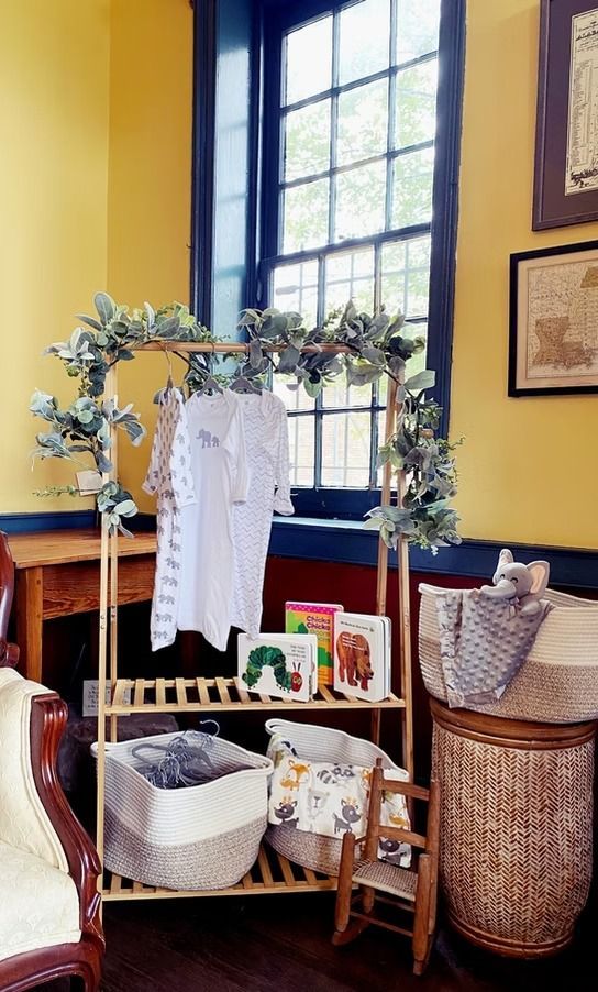 A baby clothing rack adorned with greenery stands in a yellow room next to a wicker basket with a plush toy.