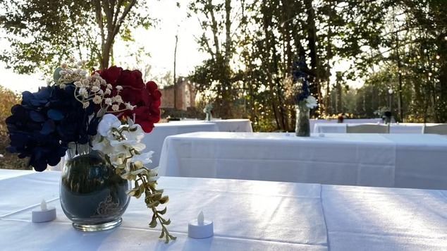 Tables with white tablecloths and patriotic red, white, and blue flower arrangements at an outdoor event during sunset.