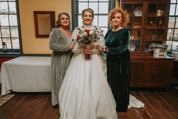 A bride in a white wedding gown holding a bouquet stands between two people in long, dark velvet dresses in a room.
