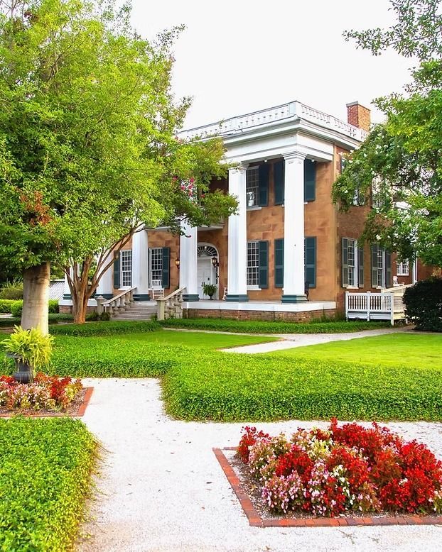 A brick antebellum-style house with tall white pillars, surrounded by green lawns and a flower bed in the foreground.