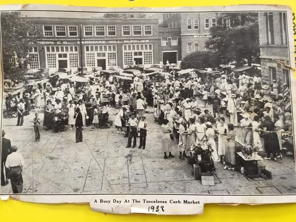 A black-and-white 1929 photo of a busy open-air street market with people gathered around stalls in front of brick buildings.