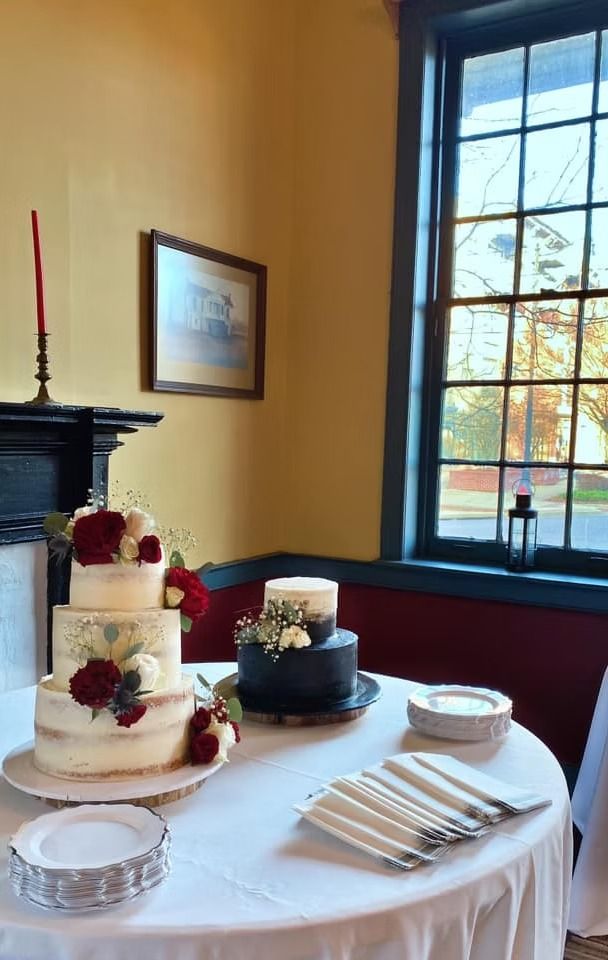 Two wedding cakes, one white and one dark, sit on a table with plates and silverware in a room with a window.