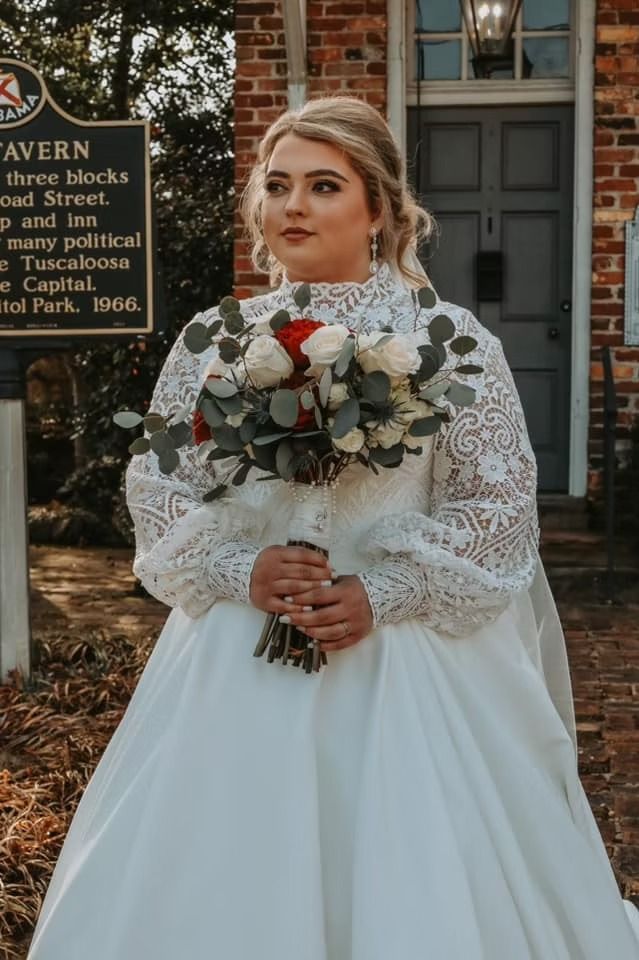 A person in a long-sleeved lace wedding dress holds a bouquet of white and red roses in front of a historic brick building.