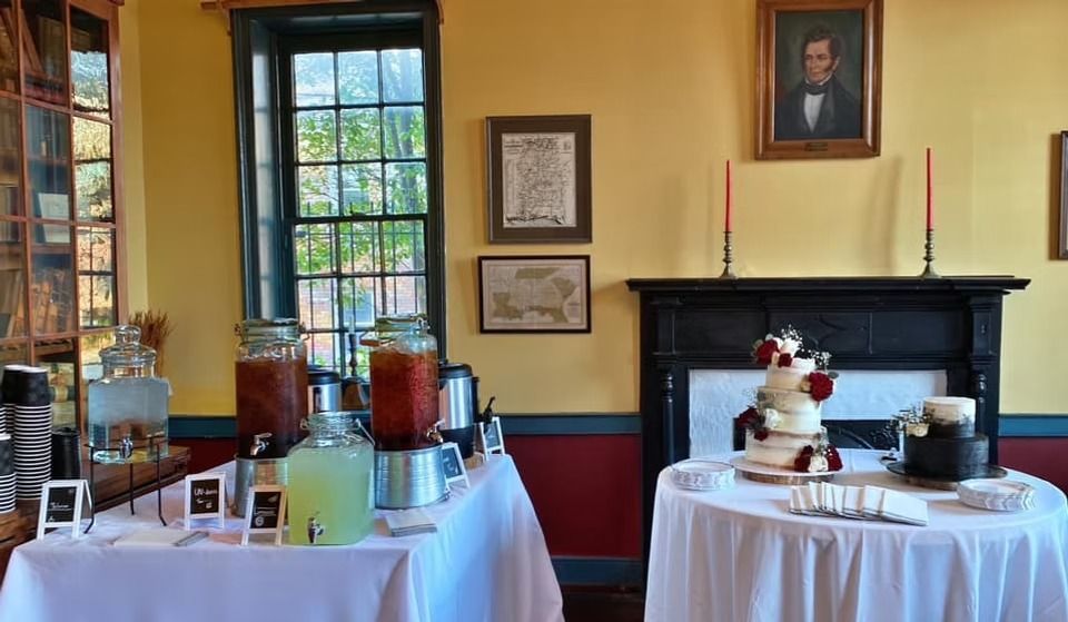 A drinks station and dessert table with two wedding cakes inside a historic room with a fireplace and portraits.