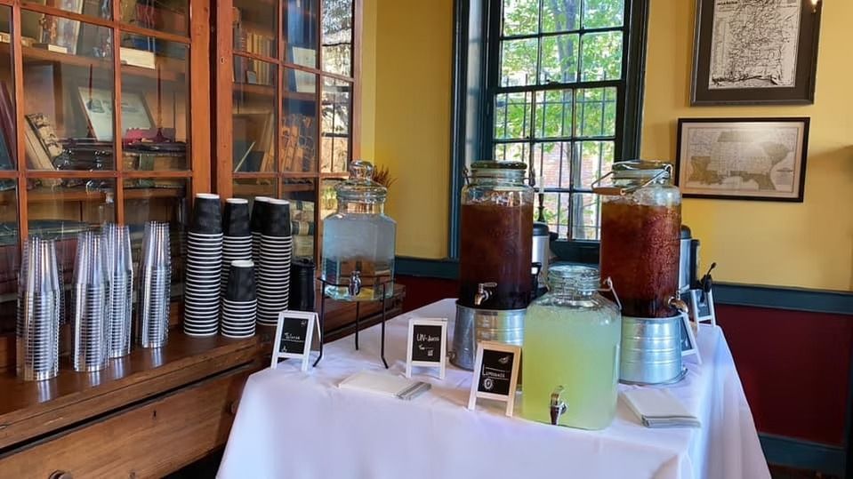 A drink station with glass beverage dispensers on a white-clothed table in a room with a wooden bookshelf and window.