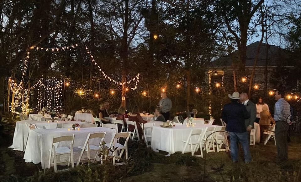 Outdoor evening event with guests at tables covered in white tablecloths, decorated with string lights among trees.