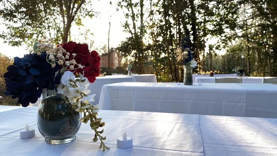 A floral centerpiece with red, white, and blue flowers on a white-covered table at an outdoor event during sunset.