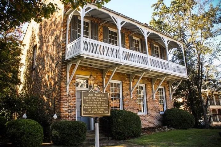 A two-story brick Old Tavern building with a wooden second-floor balcony and a historical marker sign in the front yard.