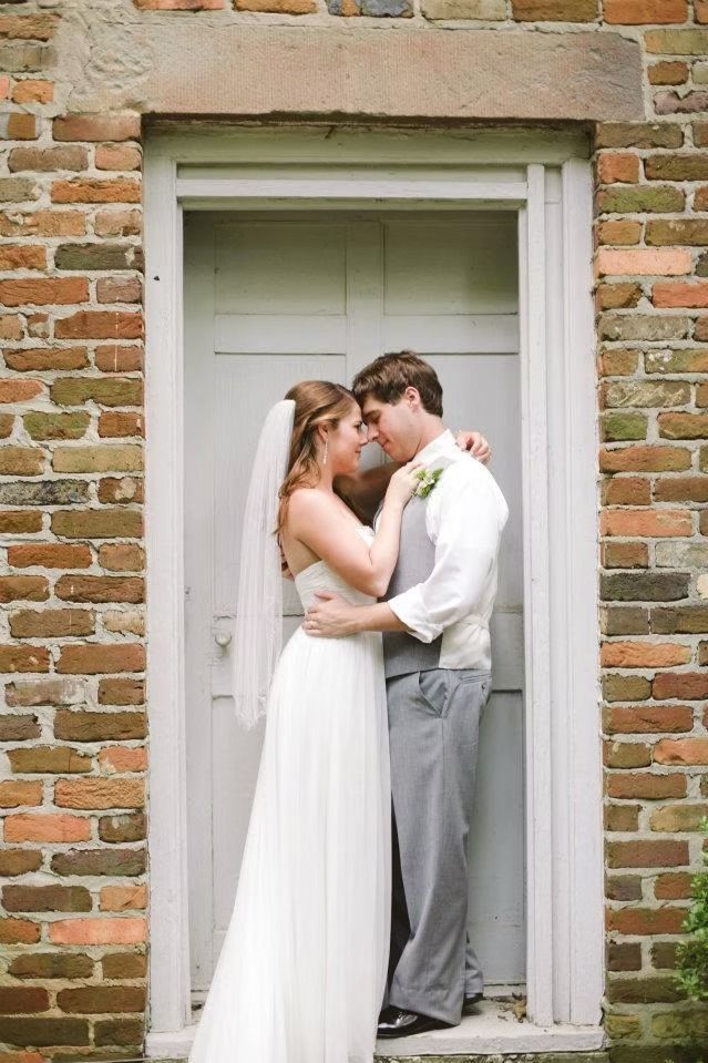 A couple in formal wedding attire embrace in a doorway framed by a rustic brick wall.