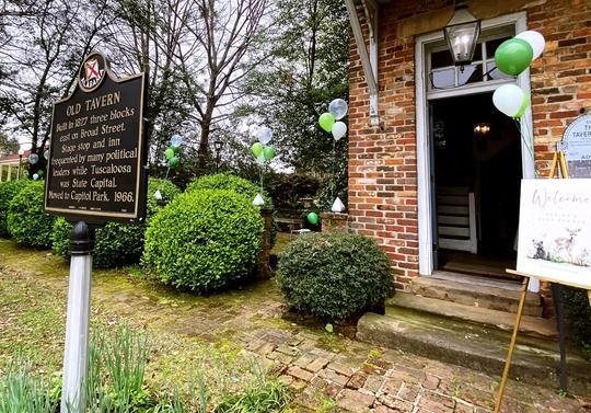 Historical brick Old Tavern building entrance with a commemorative plaque, manicured shrubs, and white and green balloons.