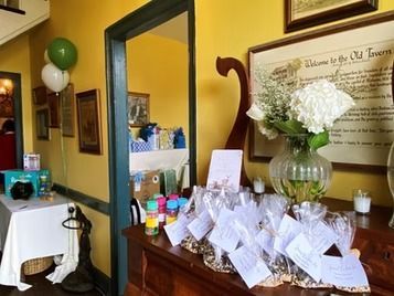 A party display with favor bags on a wooden counter, white flowers in a vase, and a table with balloons in a yellow room.
