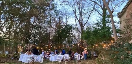 People sit at tables with white tablecloths under string lights in a wooded outdoor setting during twilight.