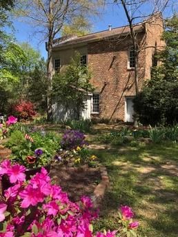A rustic brick building stands behind a garden filled with vibrant pink azaleas under a clear blue sky.