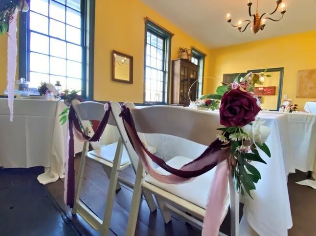 Two white folding chairs decorated with burgundy ribbons and silk flowers in a sunlit room with yellow walls.