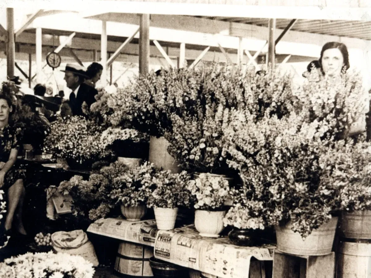 A market stall features vendors standing behind tables displaying large arrangements of flowers in pots and buckets.