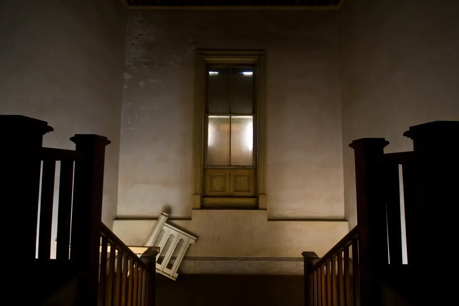 A dimly lit staircase leads to a landing with a tall, rectangular window and a discarded railing section leaning nearby.