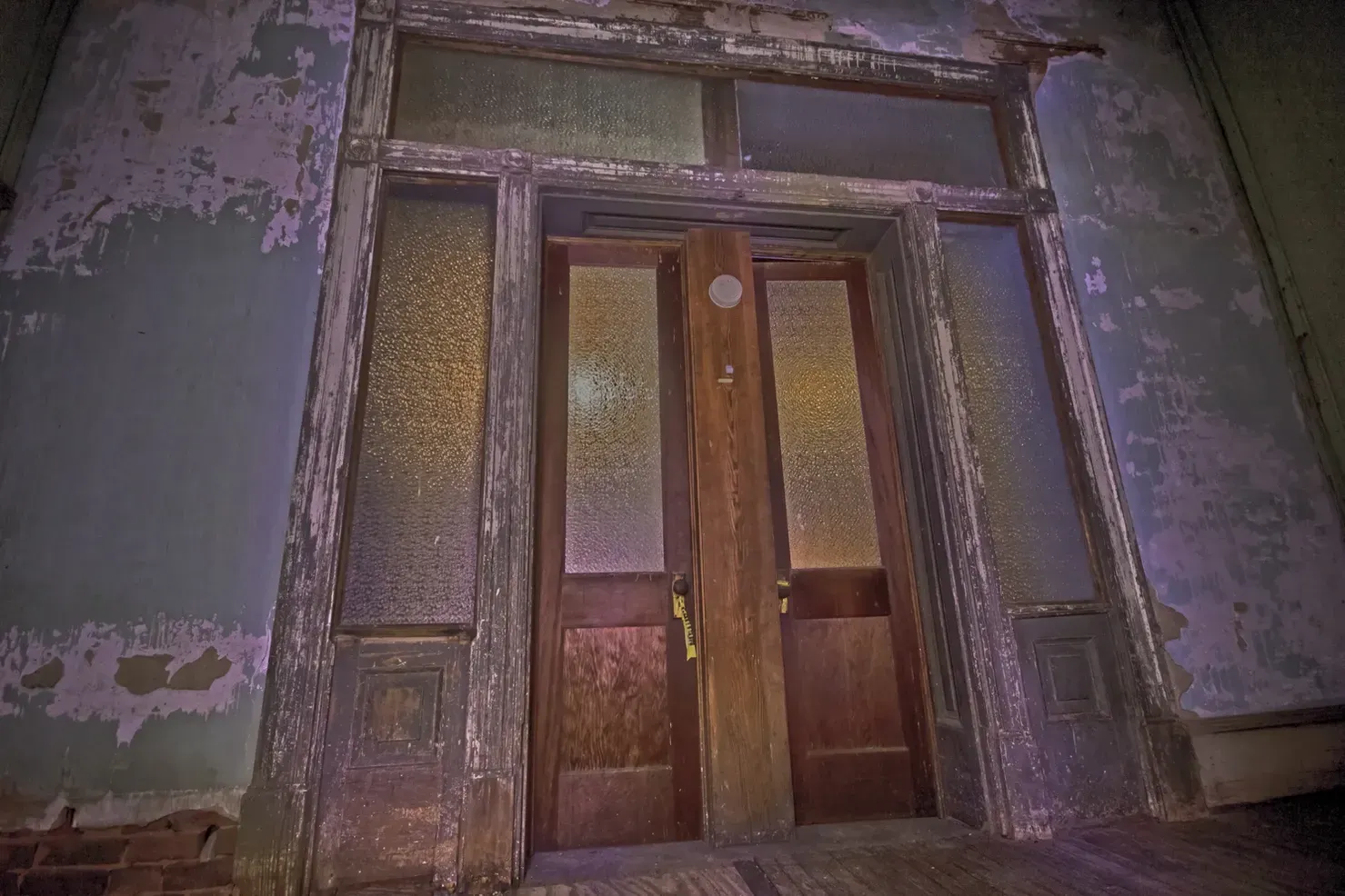 An old, weathered wooden double door with textured glass panels, set in a dilapidated wall with peeling, faded paint.