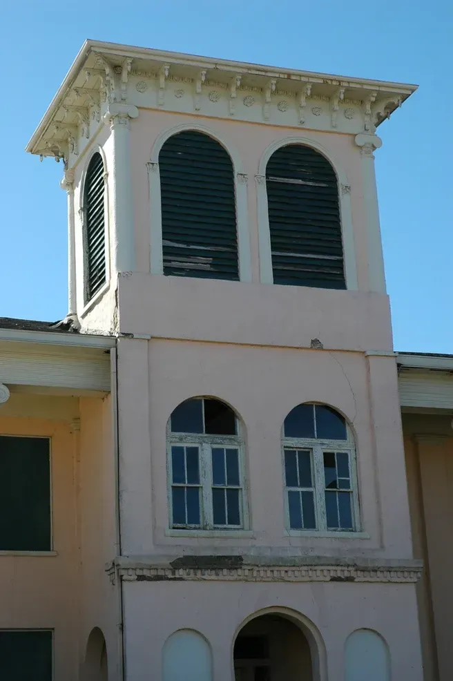 A light pink building tower with arched, shuttered windows and a decorative cornice against a clear blue sky.
