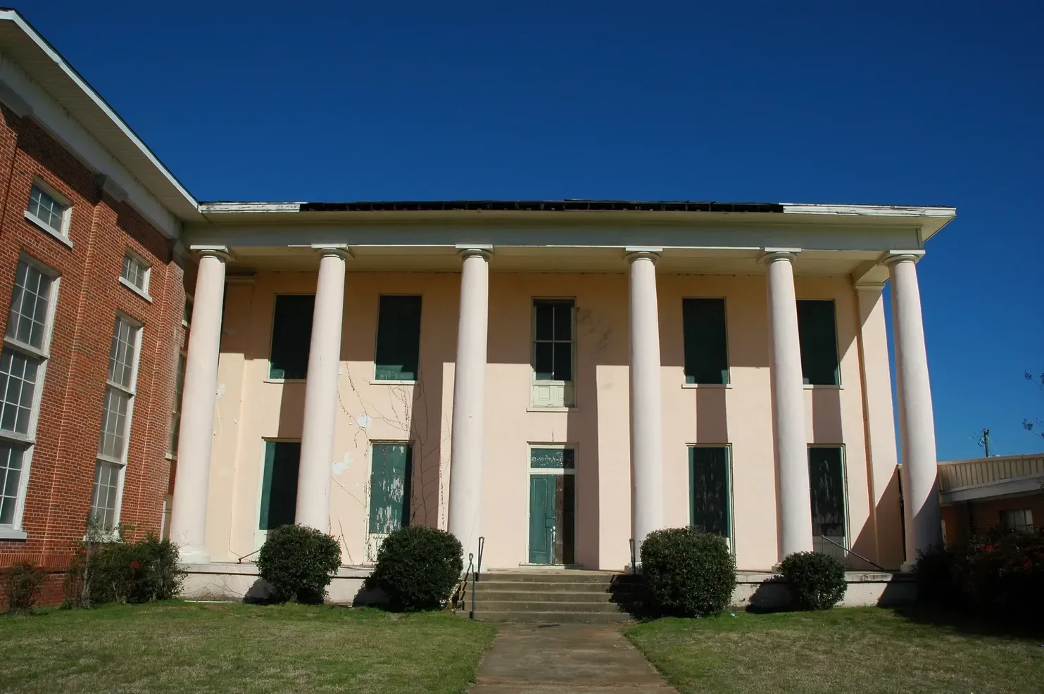 A pale pink, two-story antebellum-style house with tall white columns, dark green shutters, and a brick wing extension.
