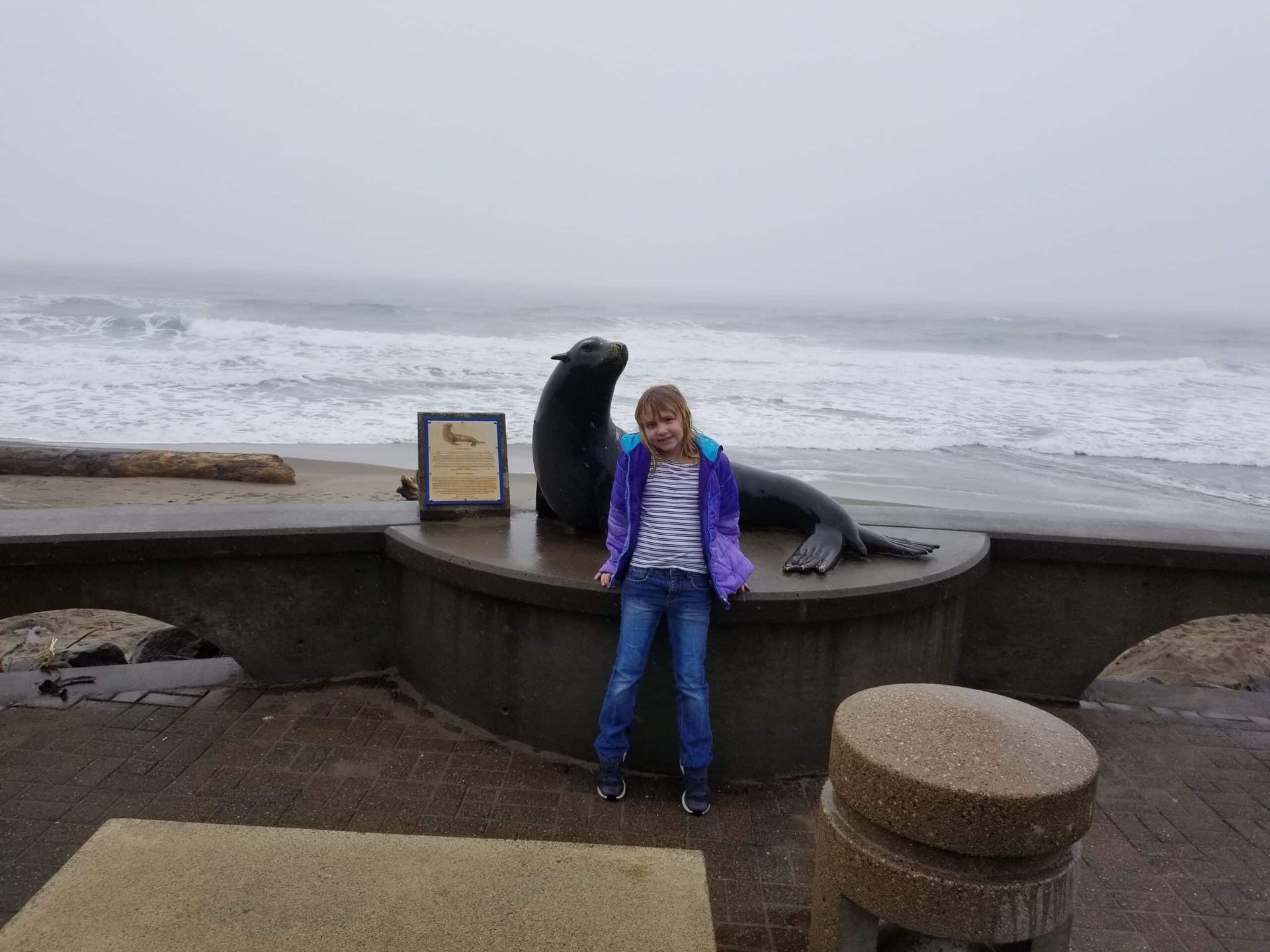 A young girl is standing next to a seal statue on the beach.