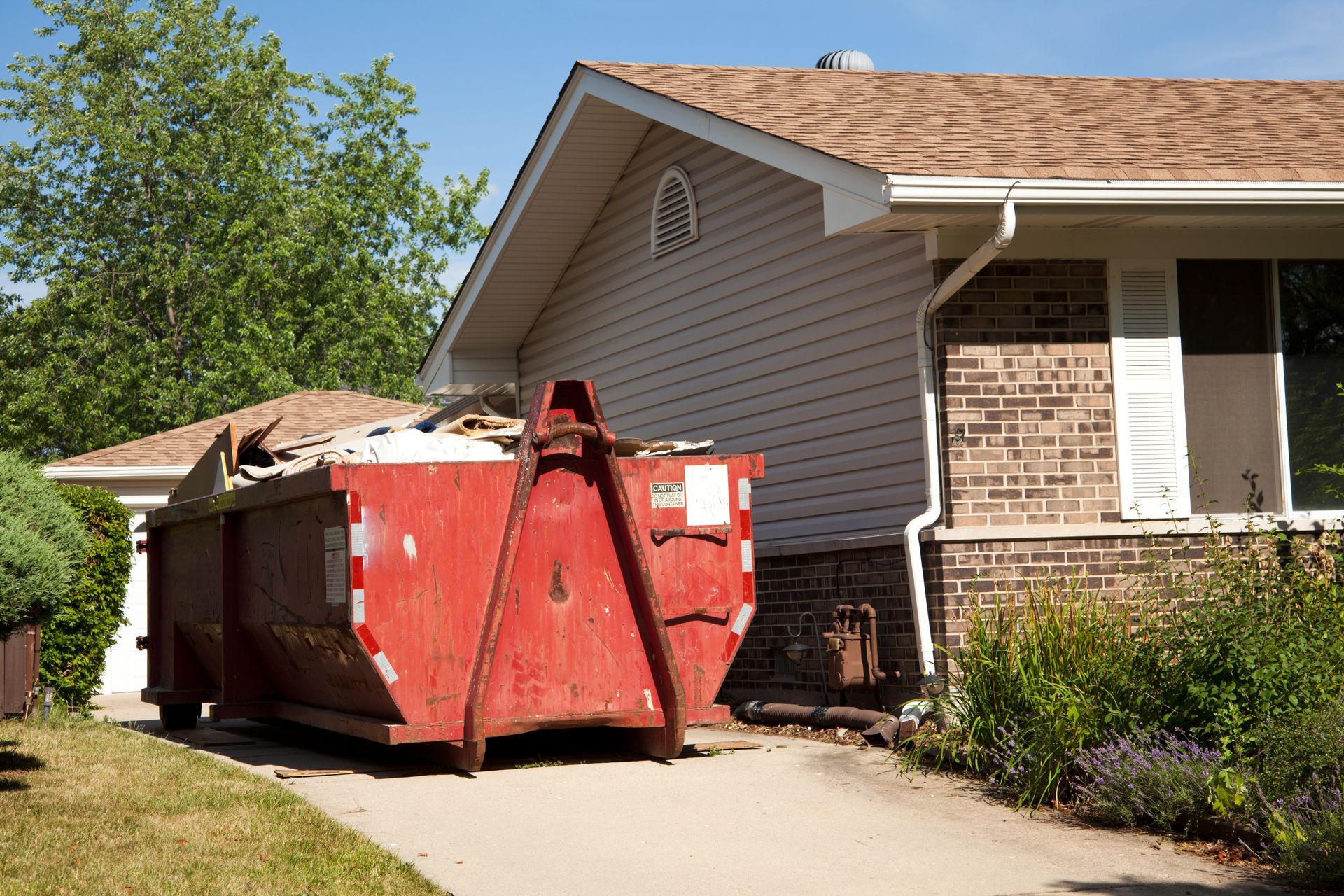 Red dumpster overflowing with debris next to a house with brick siding, on a sunny day.
