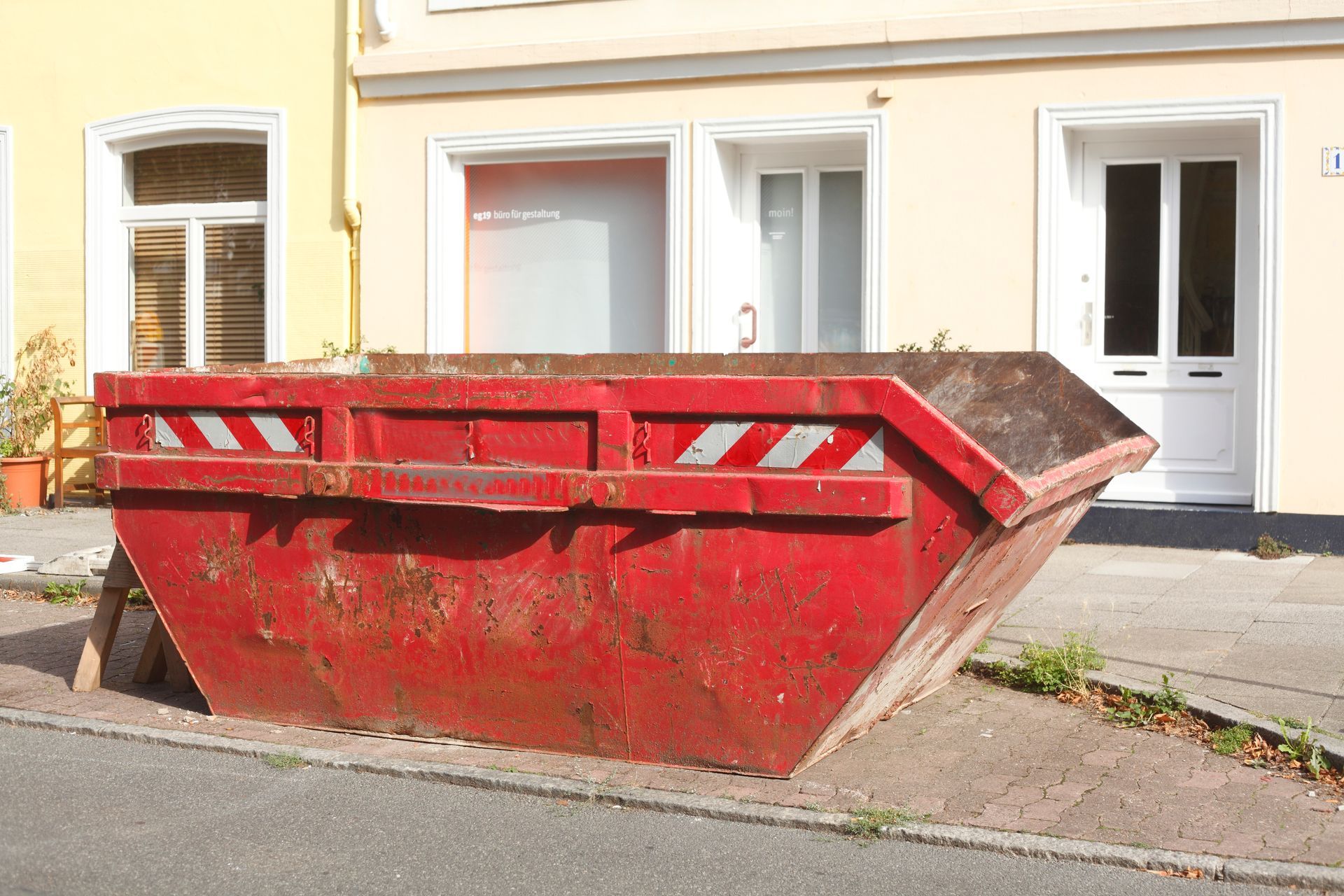 Red dumpster on a sidewalk in front of a yellow building with white doors and windows.