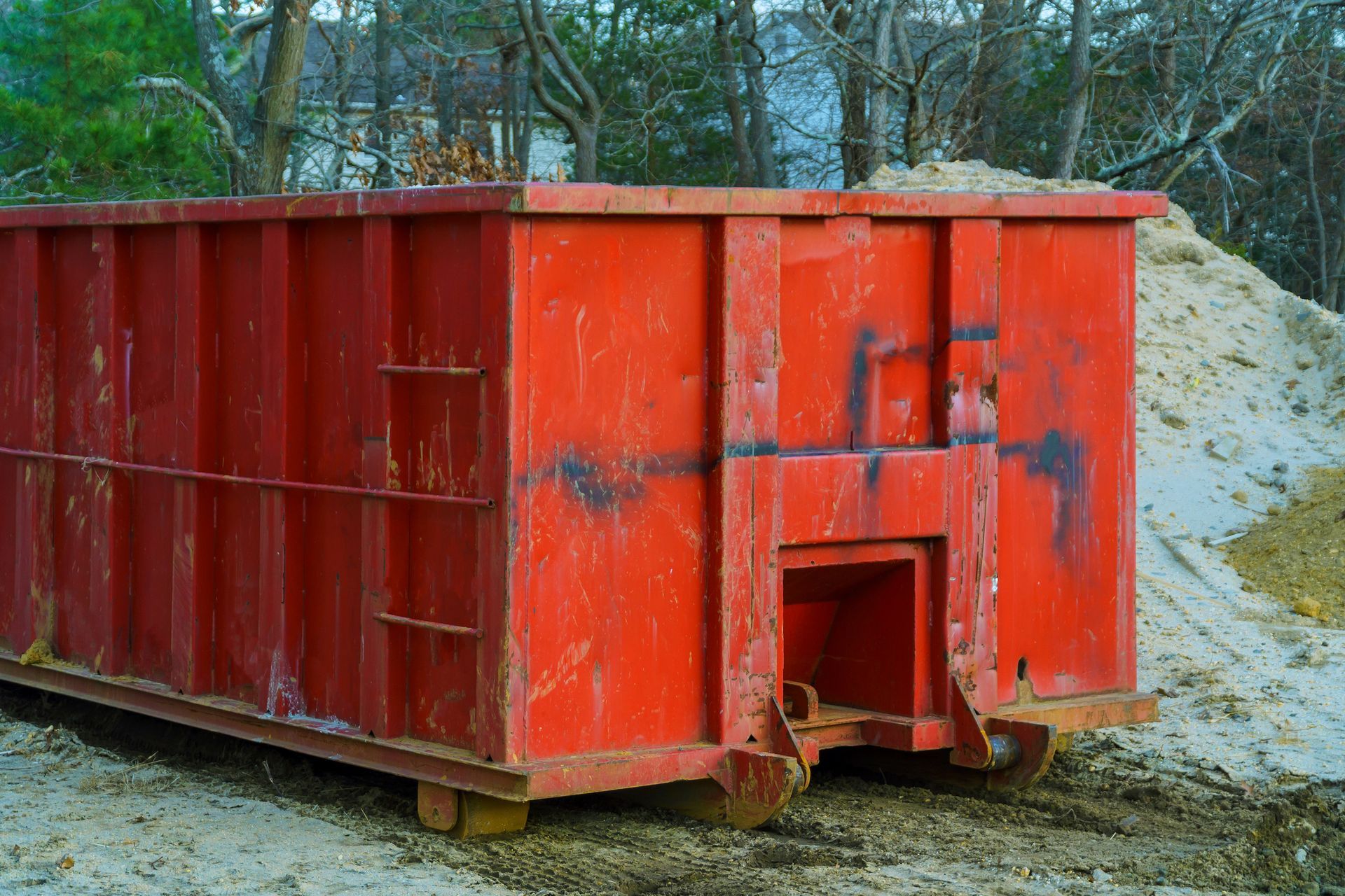 Red dumpster on wheels, parked on dirt next to a pile of sand, likely for construction debris.