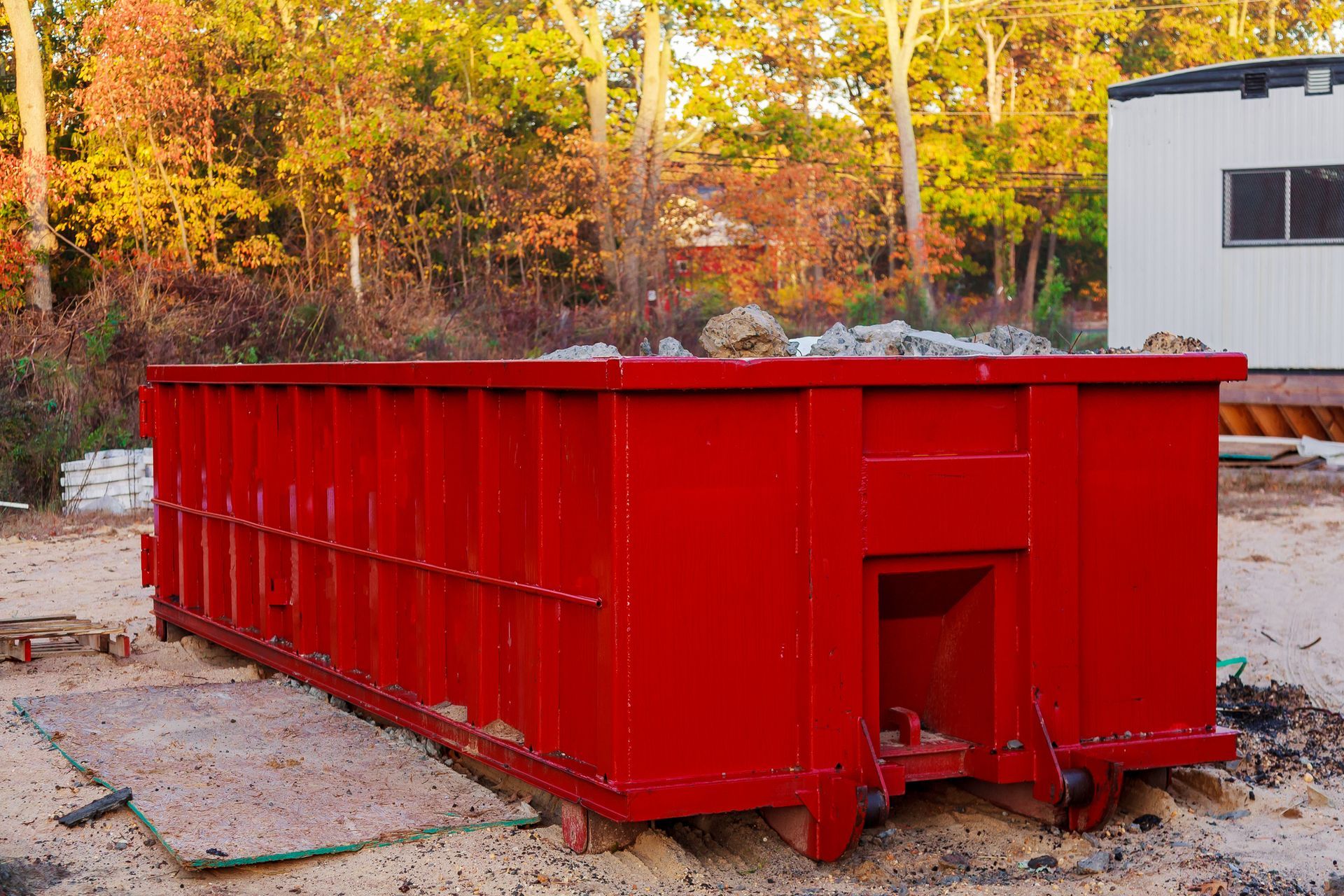 Red dumpster filled with debris, outdoors near trees and a building.