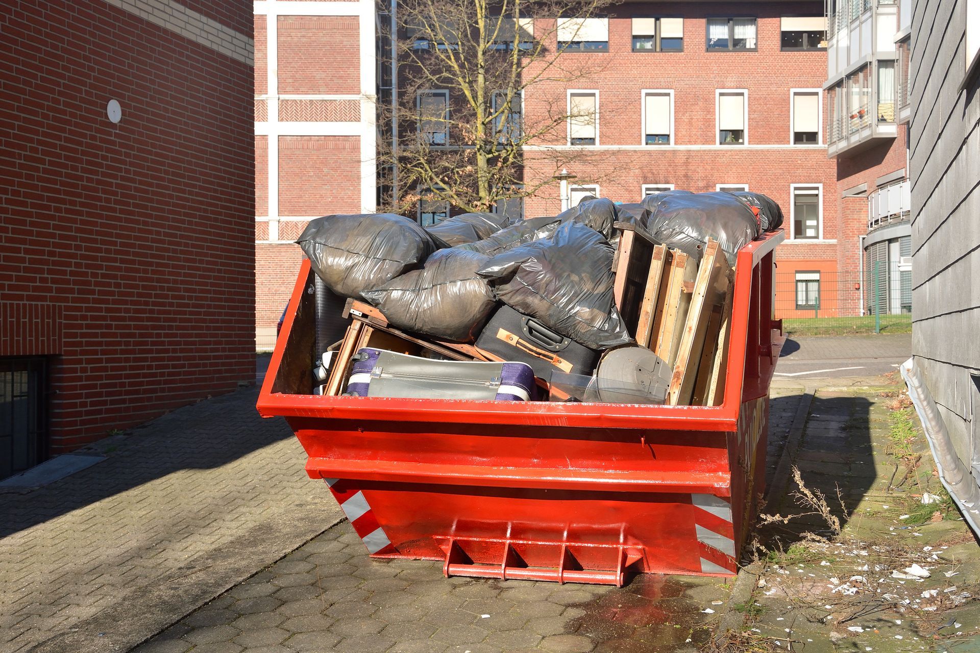 Red dumpster overflowing with trash bags and wooden pallets, in an outdoor setting next to buildings.