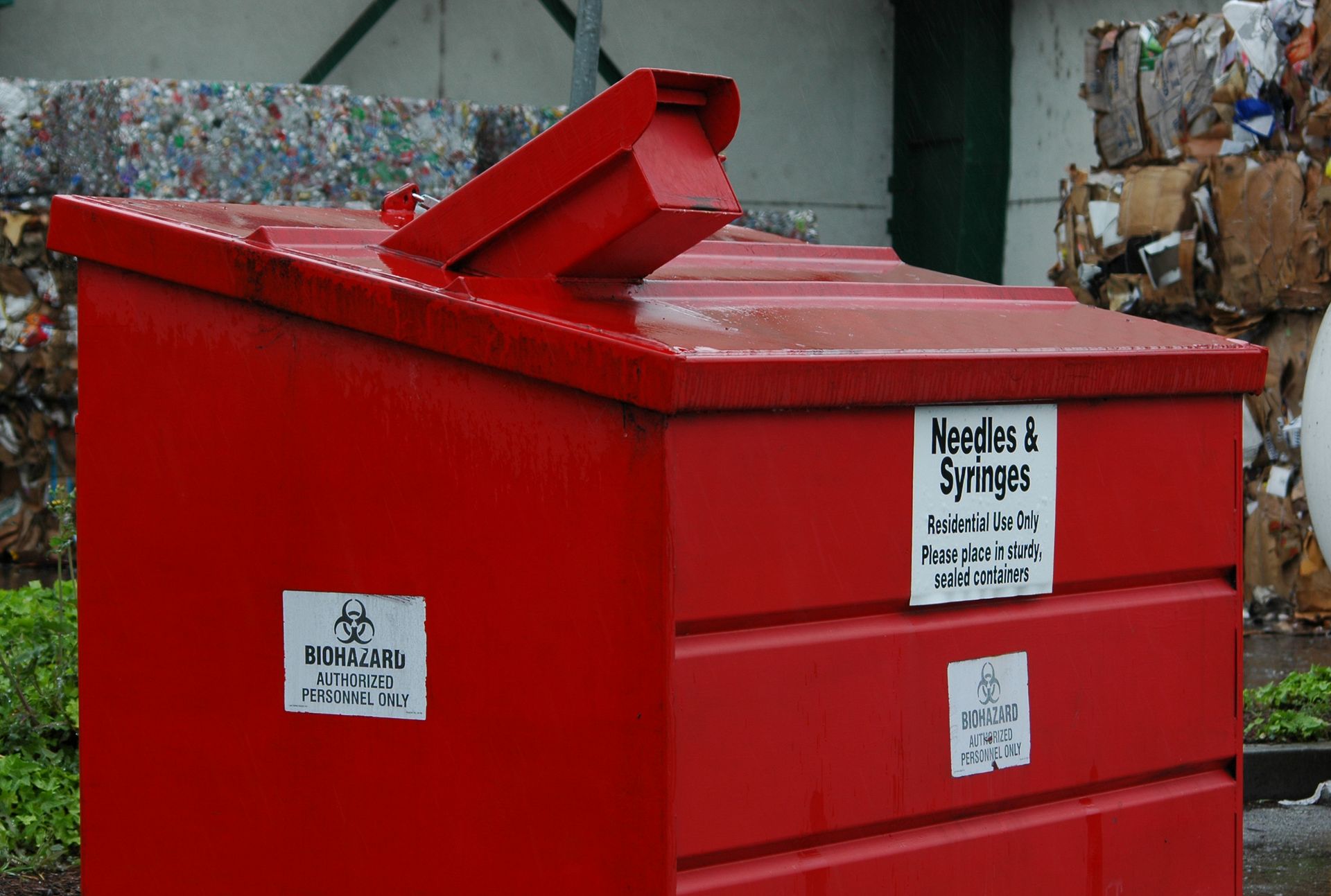 Red sharps disposal container with angled opening, biohazard labels, and text 