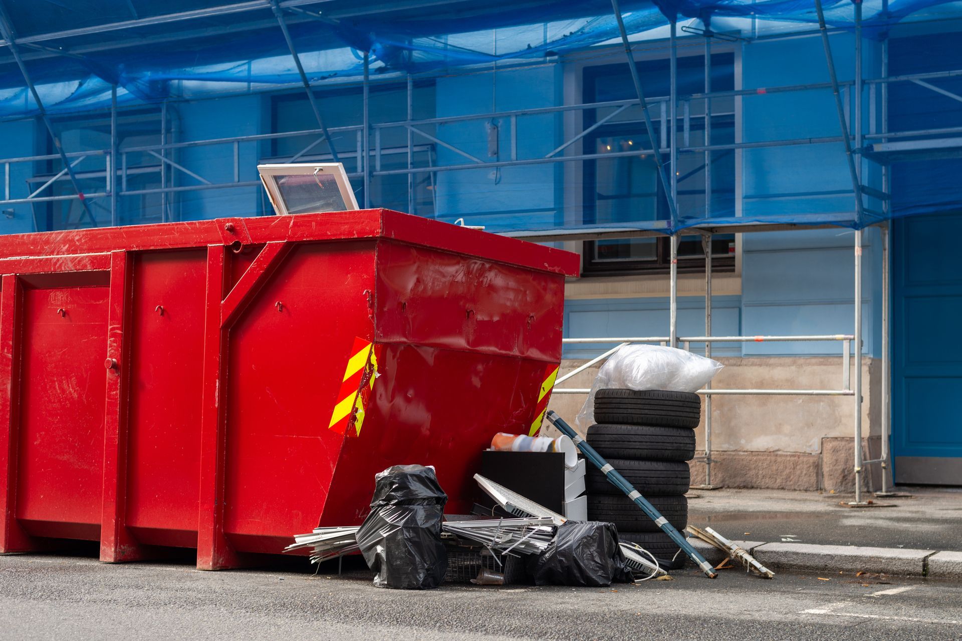 Red dumpster on city street, next to construction site, filled with trash.
