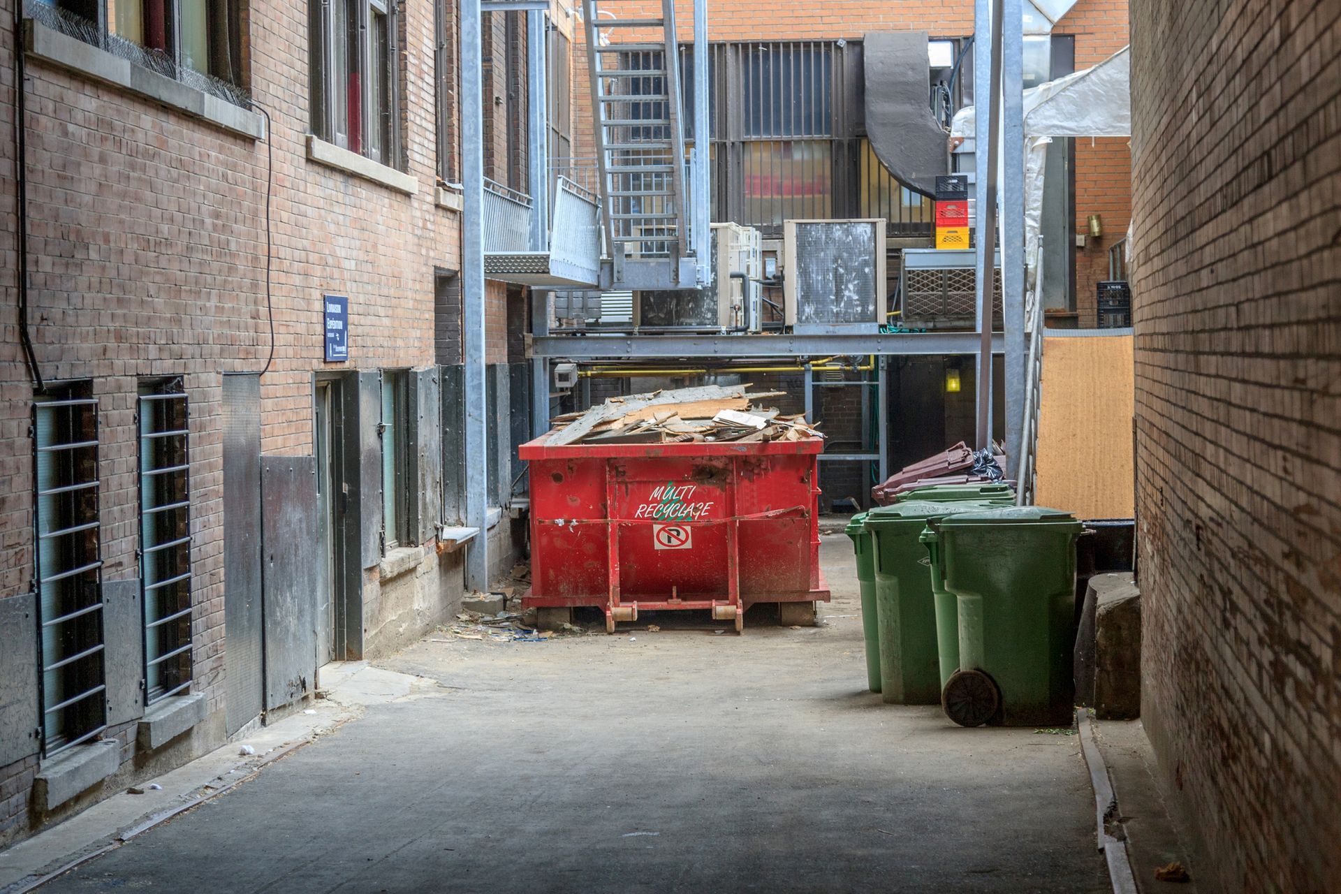Alley with red dumpster, green trash cans, and brick buildings. Metal stairs lead upward.