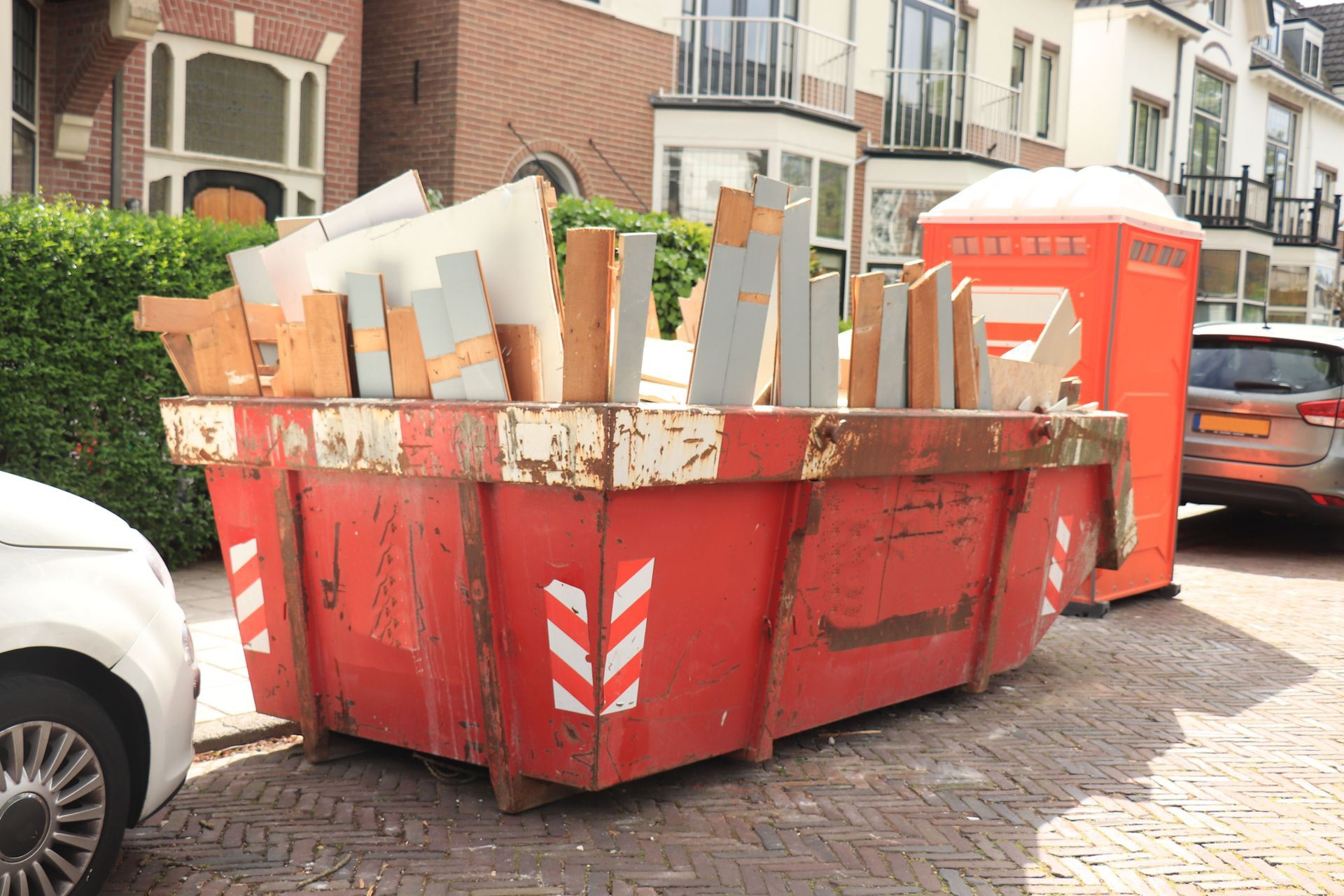 Red dumpster filled with wood scraps, next to an orange portable toilet on a brick street.