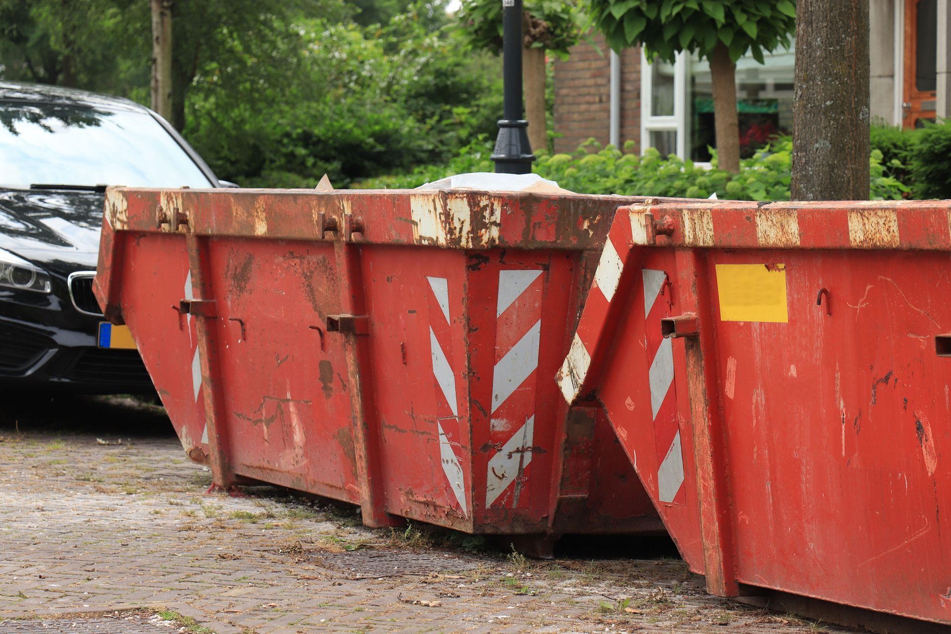 Two weathered red dumpsters on a paved surface next to a parked car, residential setting.